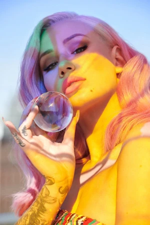 Portrait of a platinum blonde woman with colorful roots holding a clear crystal ball, light shining through the orb and creating prisms.
