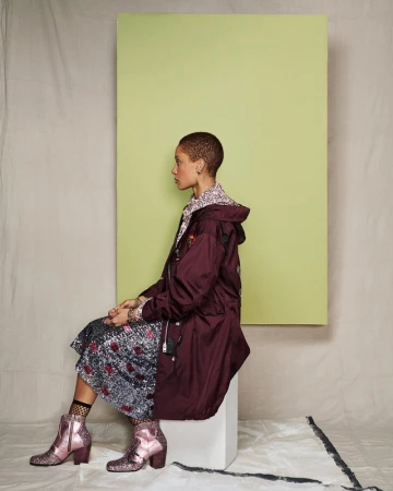 Model with a buzzcut seated in profile wearing a burgundy parka, patterned skirt, and pink glitter boots set against a green panel.