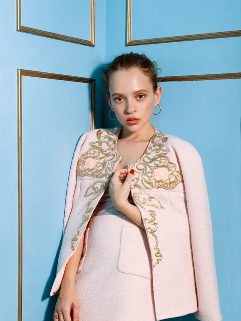 Elegant woman poses indoors against a blue wall, wearing a stylish pink jacket with gold embroidery and matching skirt.