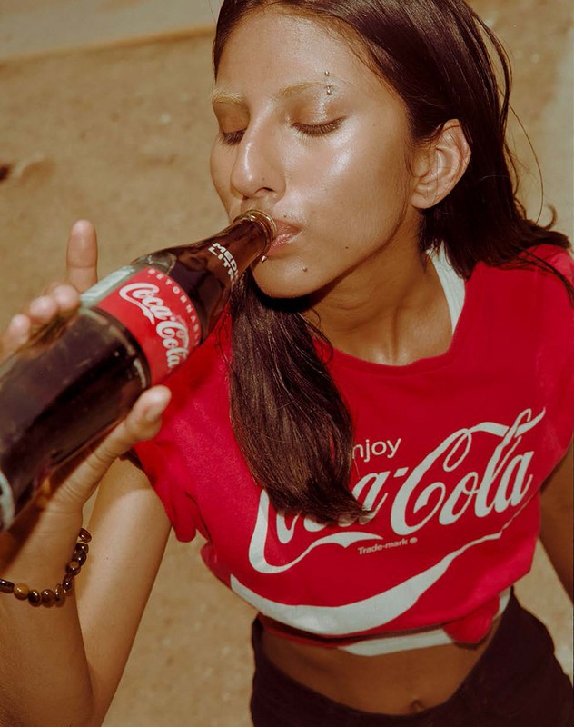 Young woman in red Coca-Cola t-shirt drinking a bottle of soda. Close-up shot with a neutral background and warm lighting.