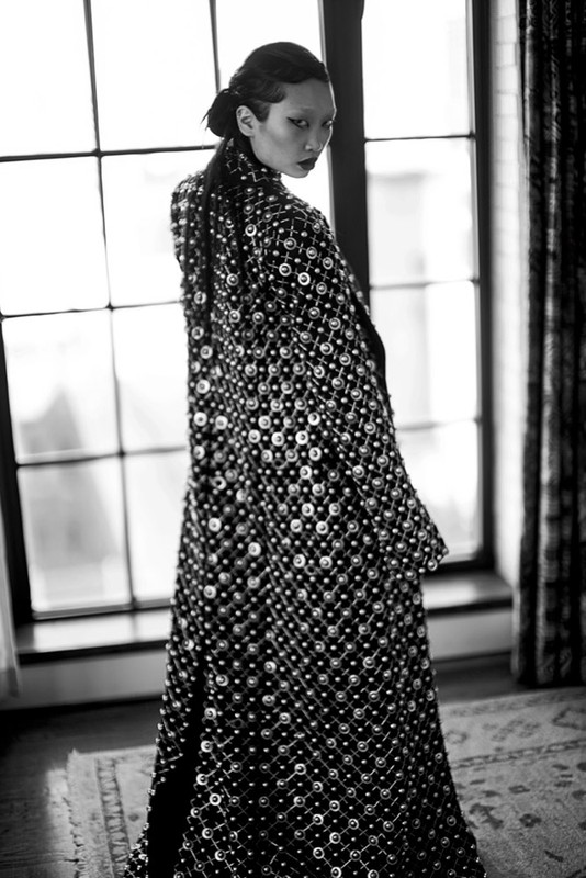 Monochrome portrait of an Asian woman in a heavily embellished coat, gazing over her shoulder toward the camera in an indoor setting.