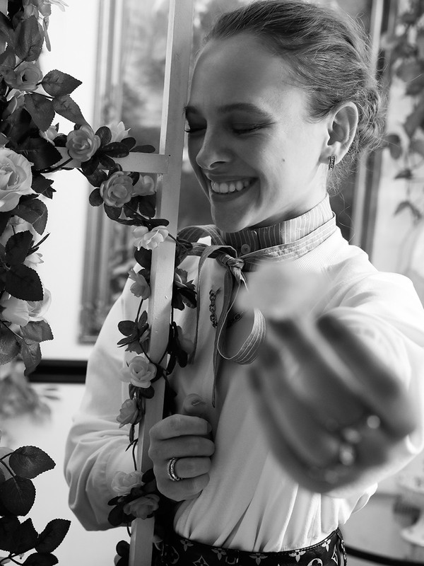 Monochrome portrait of a smiling woman in a collared shirt, holding a cross adorned with roses, in a brightly lit indoor setting.