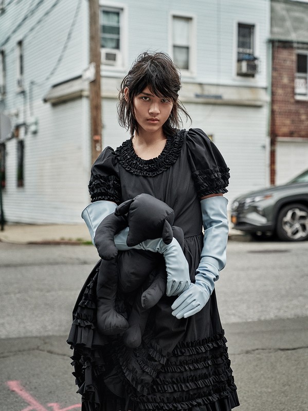 Portrait of a slender young adult woman in black dress and pale blue gloves, holding a black plush toy, on a street.