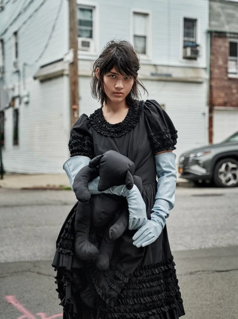Portrait of a slender young adult woman in black dress and pale blue gloves, holding a black plush toy, on a street.