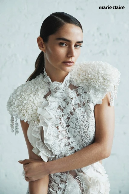 Portrait of a woman with dark hair and a serious expression, wearing a white lace top with floral detail against a white background.