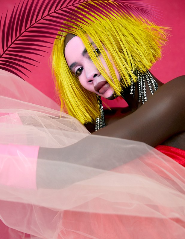 Portrait of a woman with yellow hair and large, dangly earrings, draped in pink tulle fabric against a bright pink background with palm leaves.