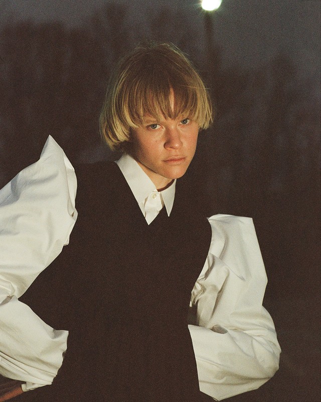 Portrait of a young adult wearing a white shirt with puffy sleeves and a black vest against a dark outdoor backdrop.