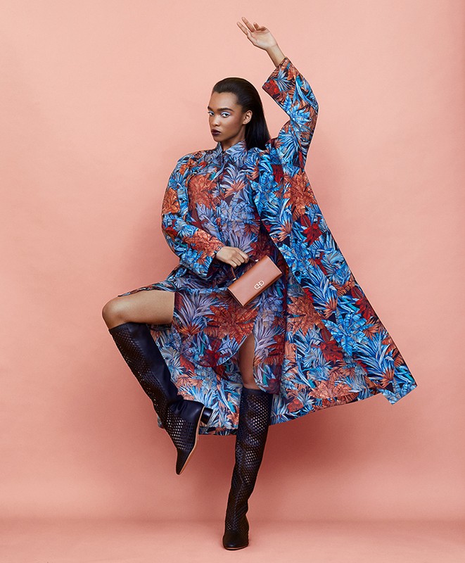 A fashion shot presents a woman in a blue floral dress and long coat, accented by tall black boots, holding a clutch on a peach background.