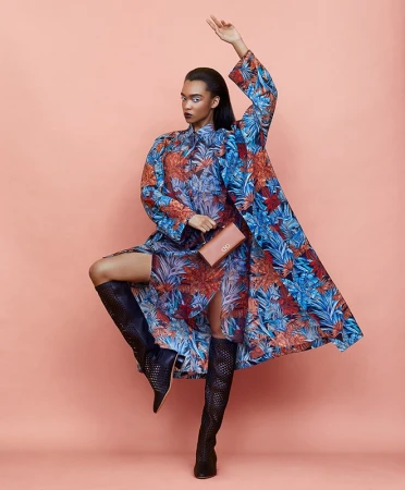 A fashion shot presents a woman in a blue floral dress and long coat, accented by tall black boots, holding a clutch on a peach background.