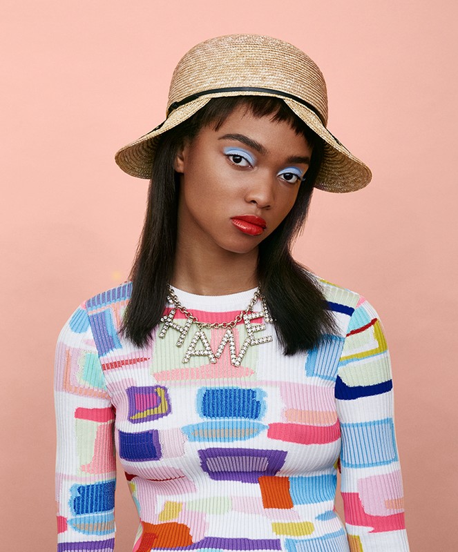 Fashion portrait of a dark-skinned woman wearing a sun hat, colorful sweater, and a HATE necklace against a peach background.
