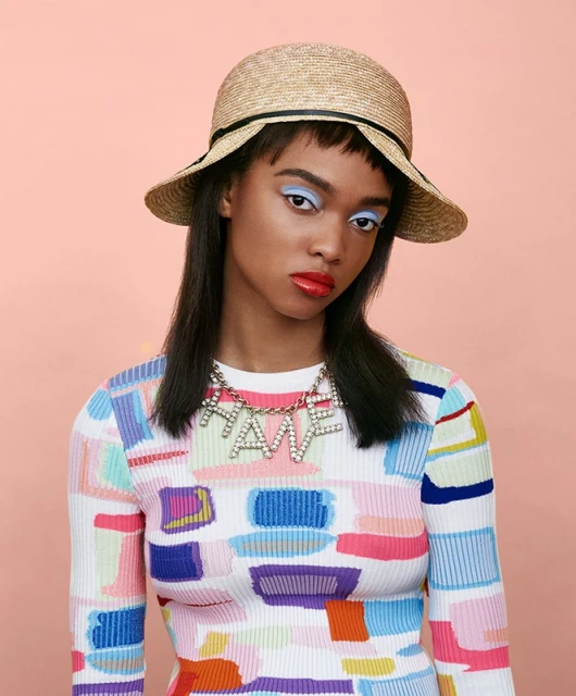 Fashion portrait of a dark-skinned woman wearing a sun hat, colorful sweater, and a HATE necklace against a peach background.
