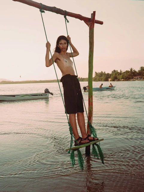 Woman in bikini top and shorts swinging on a wooden swing over water, with a boat in the distance at dusk, creating a serene scene.