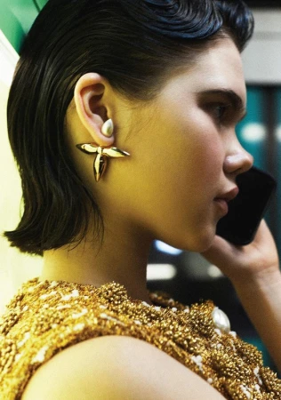 Close-up studio shot of a woman with dark, styled hair wearing a gold beaded dress and statement gold earrings, talking on a cellphone.