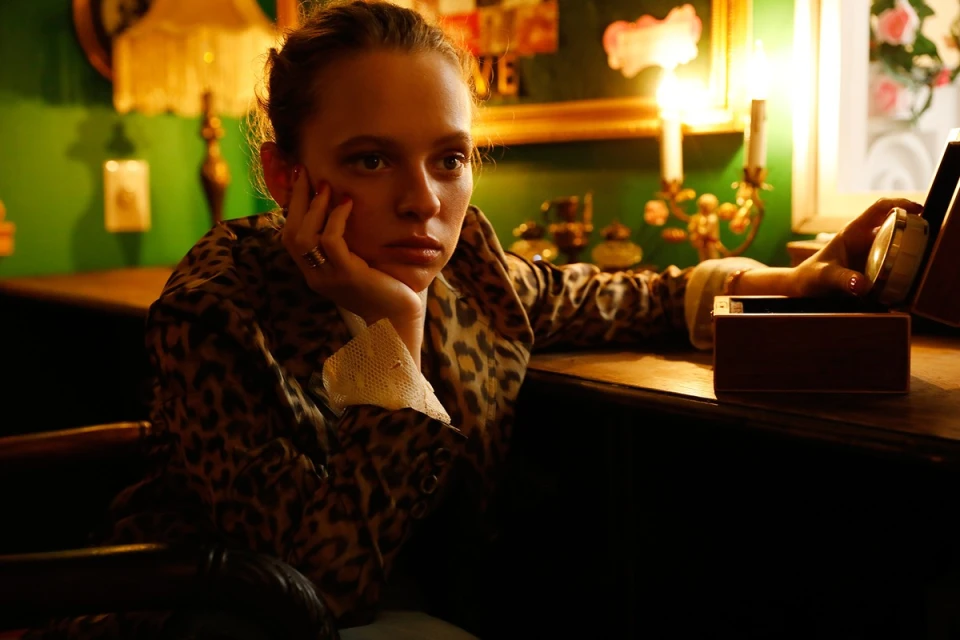 Blonde woman in animal-print jacket sits at a wooden desk indoors holding a jewelry box; moody lighting and green walls in the background.