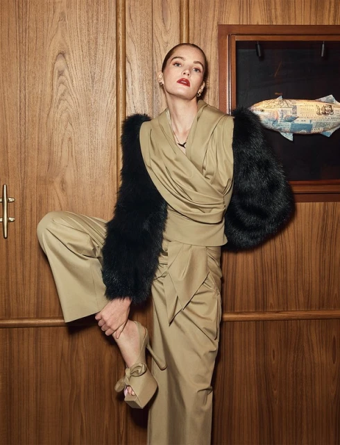 Adult woman in a tan wraparound top and wide-legged pants with black fur sleeves posing indoors against a wood-panelled wall.