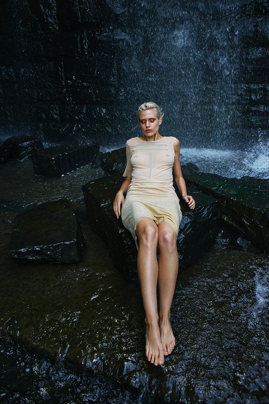Blonde woman in a beige dress seated on a dark rock in front of a rushing waterfall.