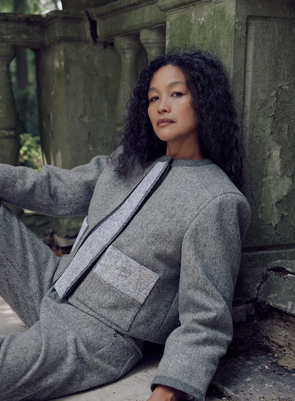 Woman with curly hair poses against weathered stone in a grey jacket and pants, in an outdoor, natural setting with green background.