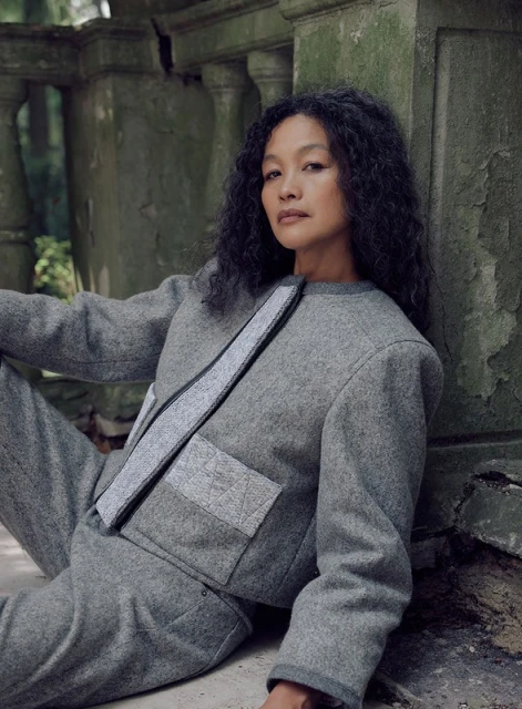 Woman with curly hair poses against weathered stone in a grey jacket and pants, in an outdoor, natural setting with green background.