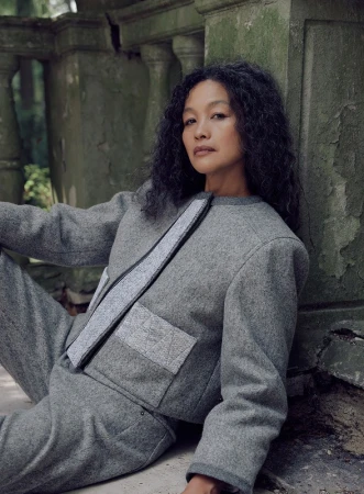 Woman with curly hair poses against weathered stone in a grey jacket and pants, in an outdoor, natural setting with green background.