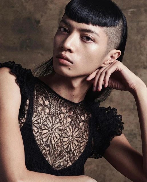 Portrait of a person with a mullet haircut and black lace top, posing indoors against a dark, mottled background in soft, diffused lighting.