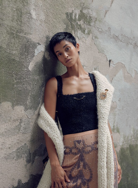 Woman with short hair poses in a black top and patterned skirt, draped with a white knit shawl against a textured concrete wall.