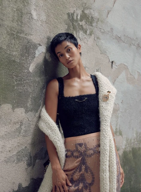 Woman with short hair poses in a black top and patterned skirt, draped with a white knit shawl against a textured concrete wall.
