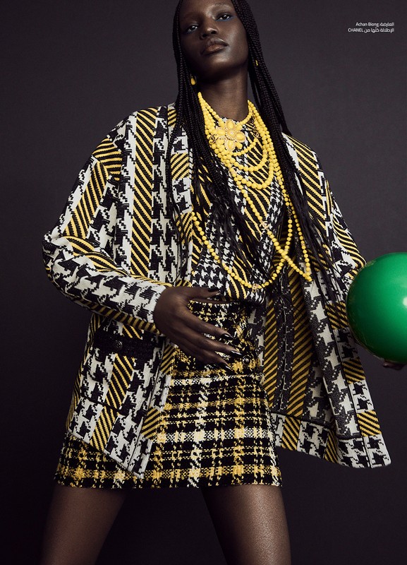 A tall African American woman wearing a yellow and black patterned jacket and skirt stands indoors, holding a green ball, jewelry visible.