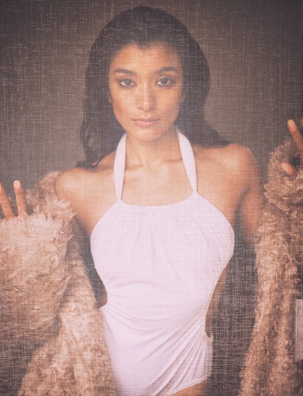 Portrait of a woman wearing a white halter swimsuit and a fur coat posing in front of a textured grey background indoors.