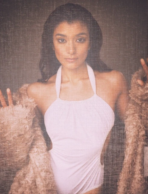Portrait of a woman wearing a white halter swimsuit and a fur coat posing in front of a textured grey background indoors.