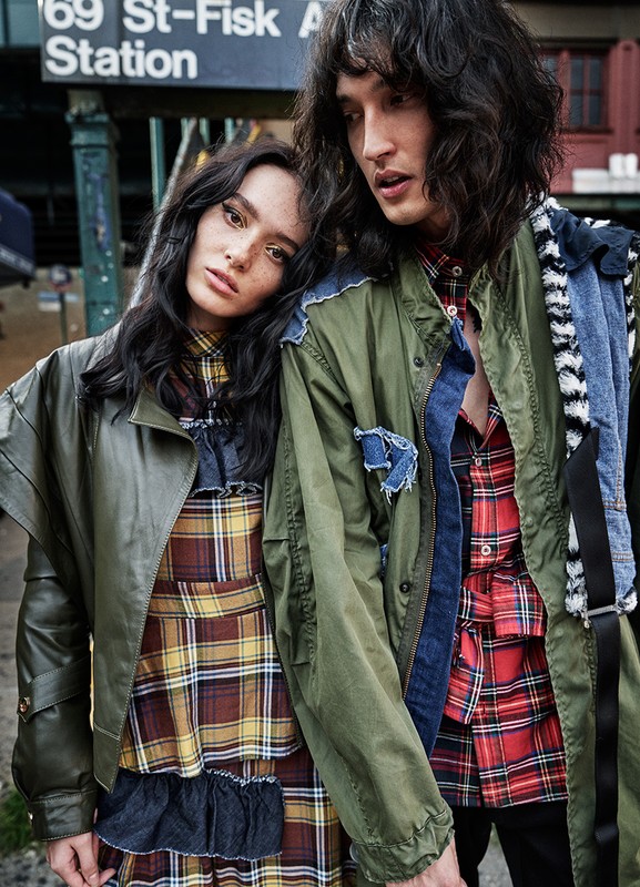 Two fashion models in stylish jackets and plaid outfits pose in front of a vintage subway station sign with dark hair and neutral faces.
