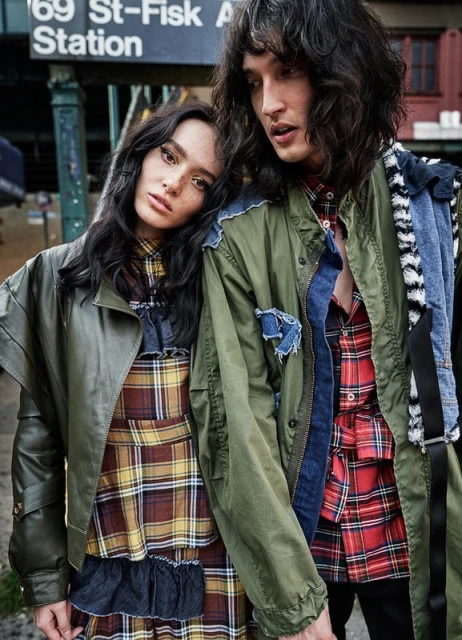 Two fashion models in stylish jackets and plaid outfits pose in front of a vintage subway station sign with dark hair and neutral faces.