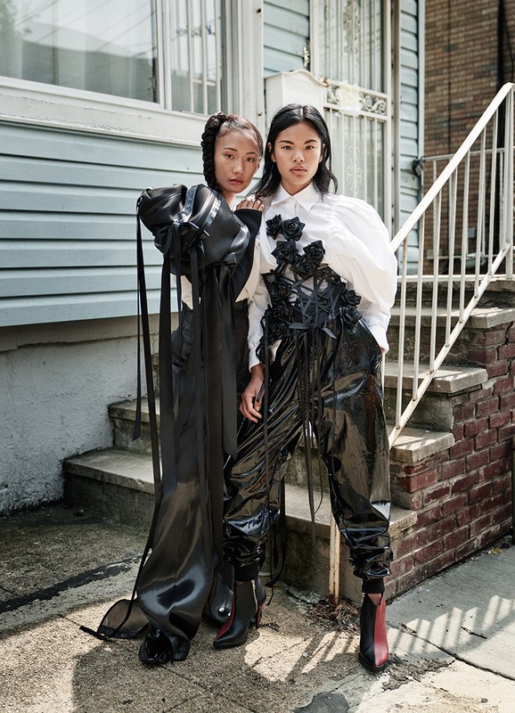 Two Asian women pose in a street setting, wearing shiny black avant-garde outfits with contrasting white blouses and textured black embellishments.