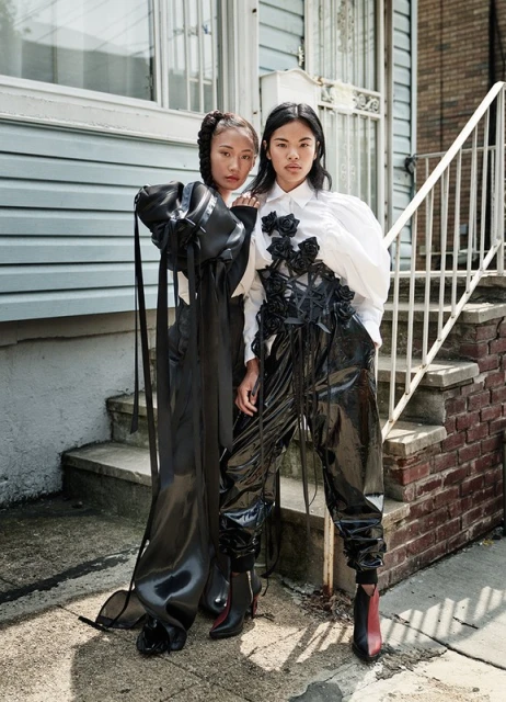 Two Asian women pose in a street setting, wearing shiny black avant-garde outfits with contrasting white blouses and textured black embellishments.