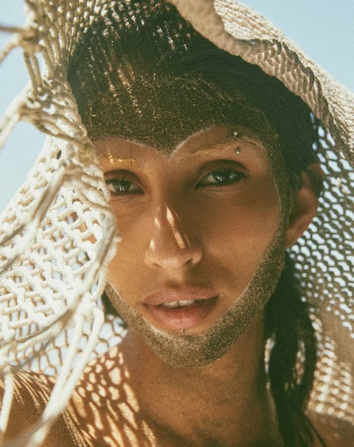 Close-up of a person with dark hair and tan skin, partially obscured by a tan woven net, illuminated by natural light.