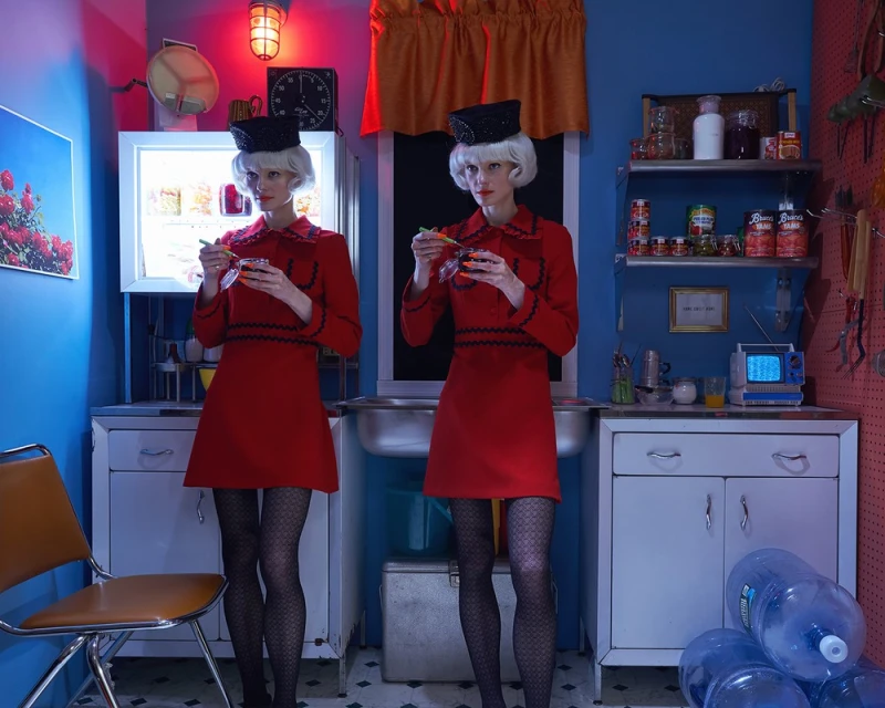 Two women with white hair and matching red dresses stand in a blue kitchen, eating from bowls under bright lighting and vintage decor.