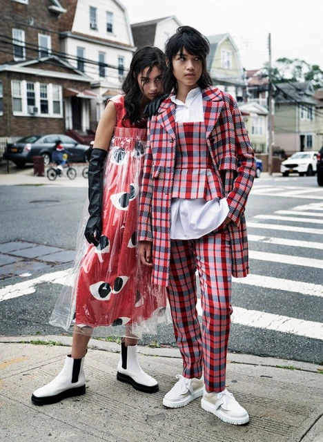 Two fashion models pose in an urban street. One wears a red dress with eye-print design, while the other sports a plaid coat and pants.