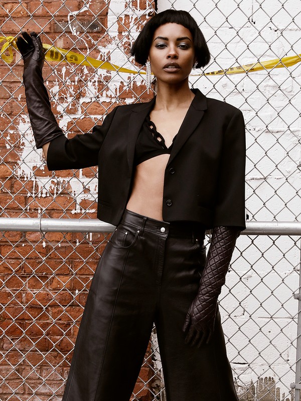 Attractive African-American woman poses in wide-leg leather pants, bra top, and a short jacket, with brick wall and chain-link fence.