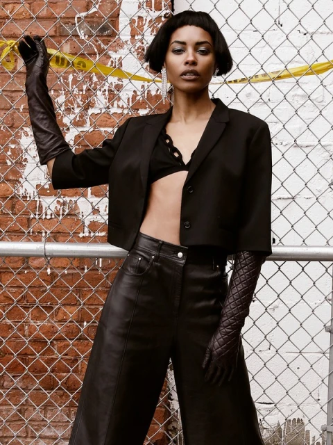 Attractive African-American woman poses in wide-leg leather pants, bra top, and a short jacket, with brick wall and chain-link fence.