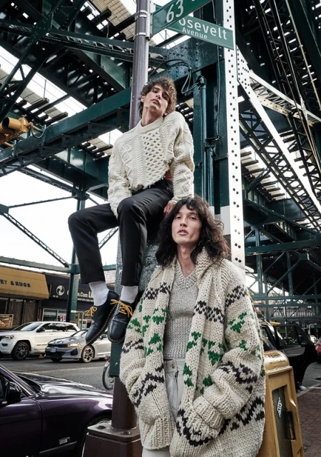 Two adult males showcasing knitwear fashion in an urban outdoor setting with cars, street signs, and elevated train tracks.