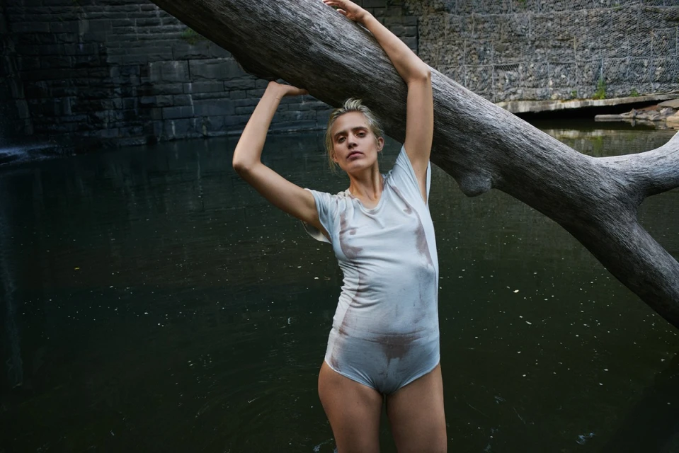 Adult woman in a wet white top and briefs poses with a large tree branch in a natural outdoor setting near a body of water.