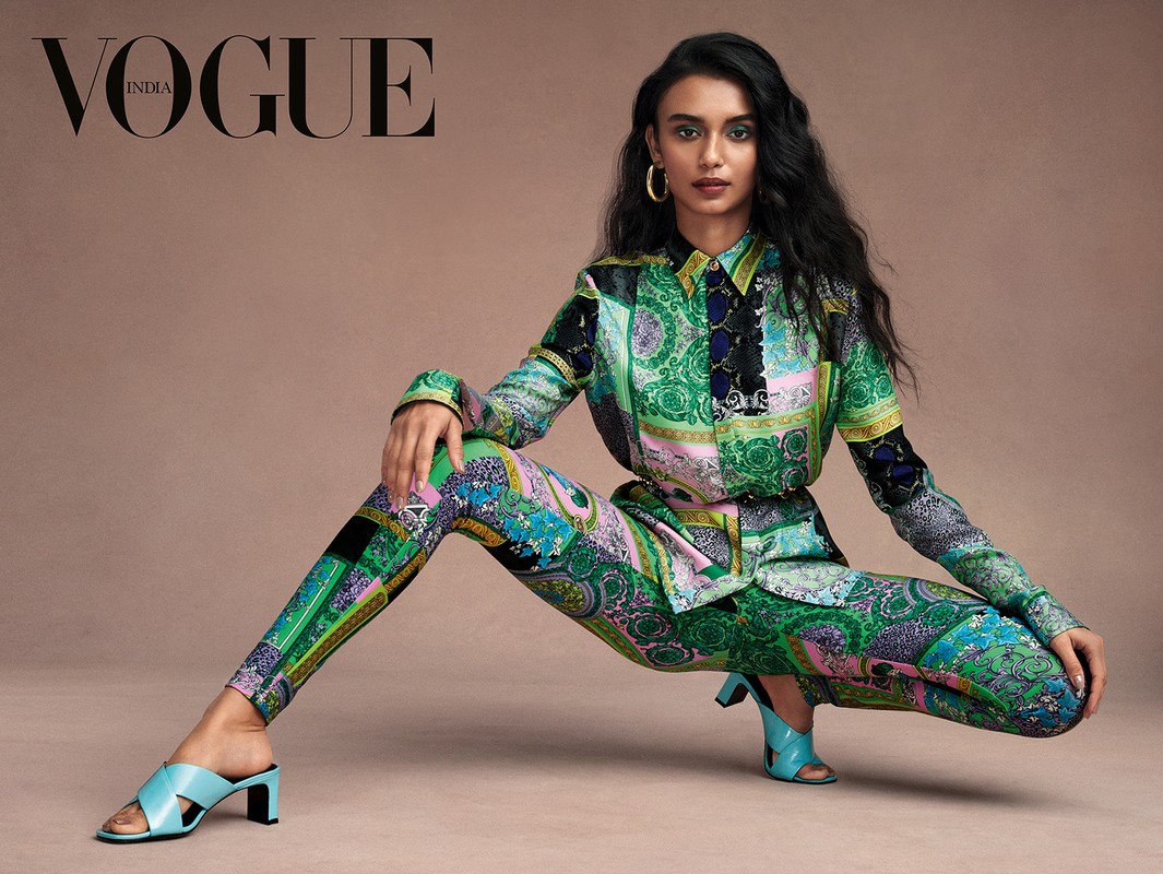 A South Asian fashion model poses in a studio wearing a patterned shirt, pants, and heels, accessorized with hoop earrings, against a neutral backdrop.