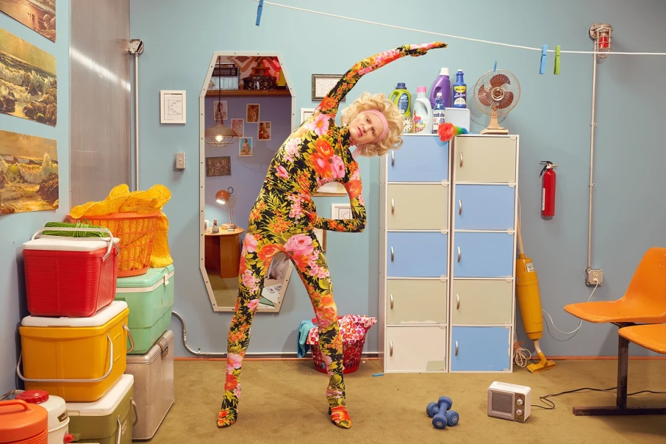 Woman in a floral bodysuit stretches in a retro laundry room with colorful coolers, cleaning supplies, and vintage decor. 