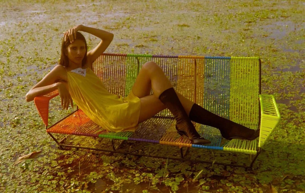 A slender woman in a yellow slip dress and boots reclines on a rainbow-colored woven bench surrounded by green plants and reflective water.