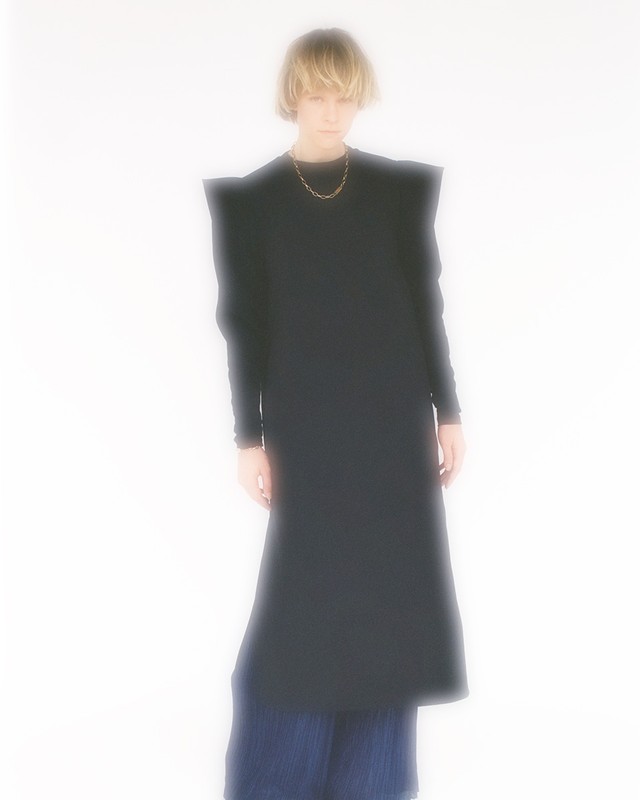 Adult wearing a long black dress and pleated blue pants stands in a bright white indoor studio, posing for a fashion editorial shot.