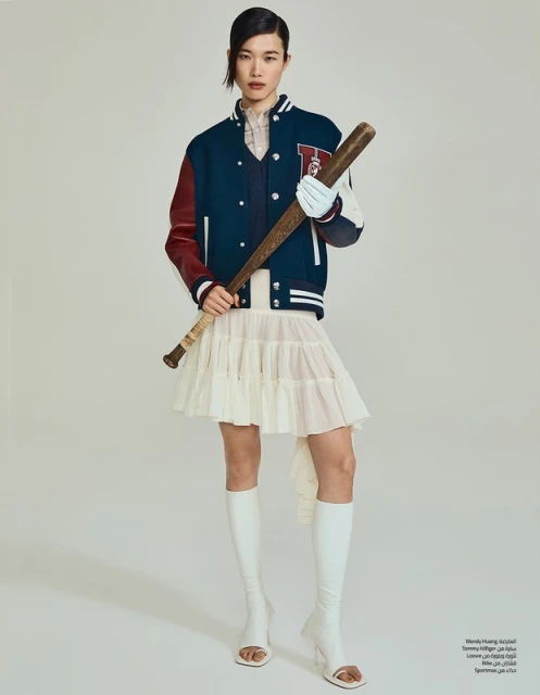 A young Asian woman strikes a pose in a fashion shot, sporting a baseball jacket, skirt, boots, and clutching a baseball bat in studio lighting.