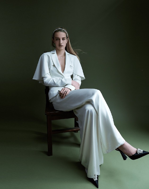 Fashion portrait of a woman in a white suit sitting on a chair against an olive-green backdrop, showcasing elegant attire and poised style.
