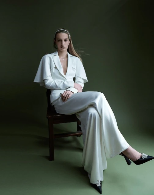 Fashion portrait of a woman in a white suit sitting on a chair against an olive-green backdrop, showcasing elegant attire and poised style.