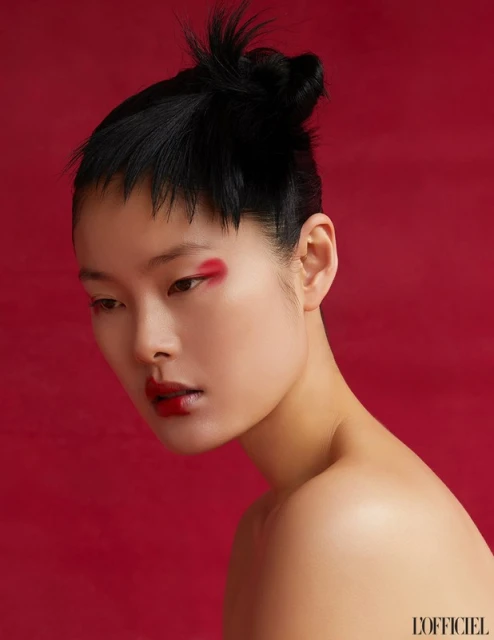 Portrait of a woman with a unique hairstyle and vibrant red makeup on her eyes and lips, set against a matching solid red background.