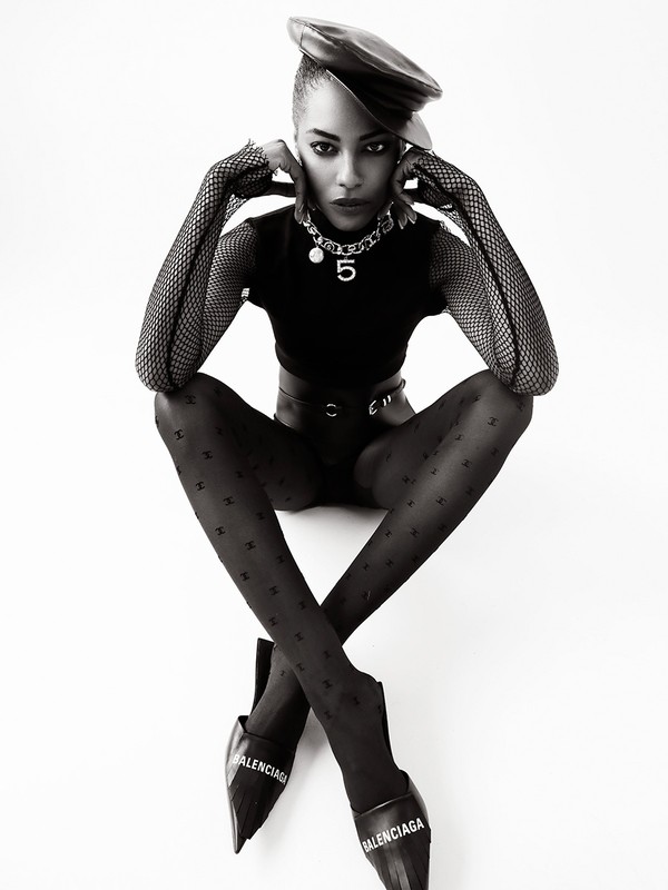 Black and white fashion portrait of an adult woman in patterned tights, a leather beret, a top and Balenciaga footwear in a studio.