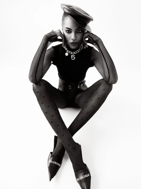Black and white fashion portrait of an adult woman in patterned tights, a leather beret, a top and Balenciaga footwear in a studio.
